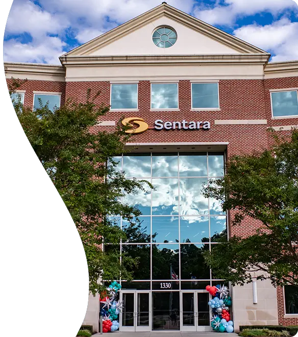Exterior view of a brick building with large reflective glass windows and the "Sentara" logo displayed above the entrance; colorful balloon decorations flank the doorway, and trees partially frame the scene under a partly cloudy sky.