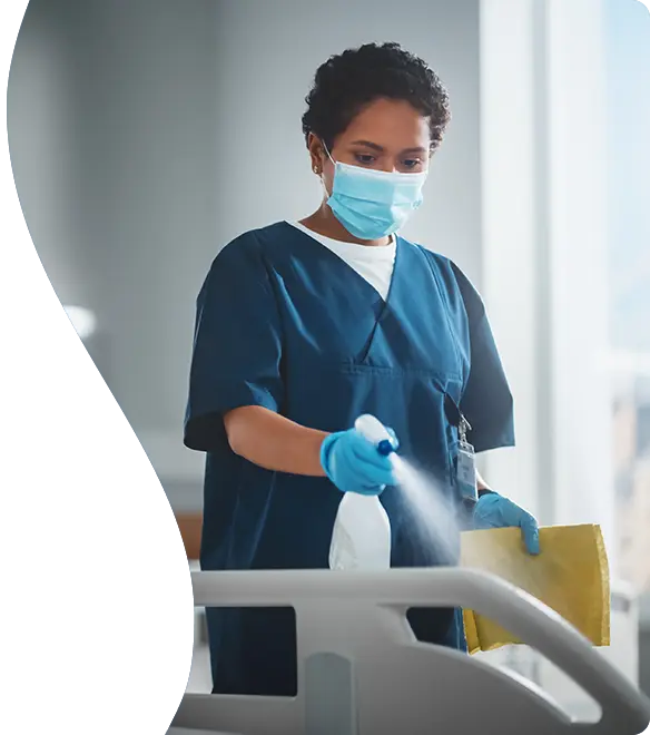 A healthcare worker wearing scrubs, gloves, and a face mask disinfects a hospital bed with a spray bottle and cleaning cloth in a brightly lit medical facility.