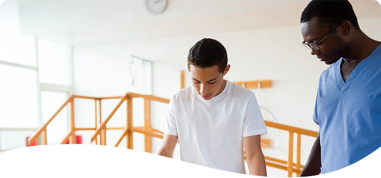 A young man wearing a medical walking boot practices walking with the support of parallel bars while a healthcare professional in scrubs closely assists him in a physical therapy setting.