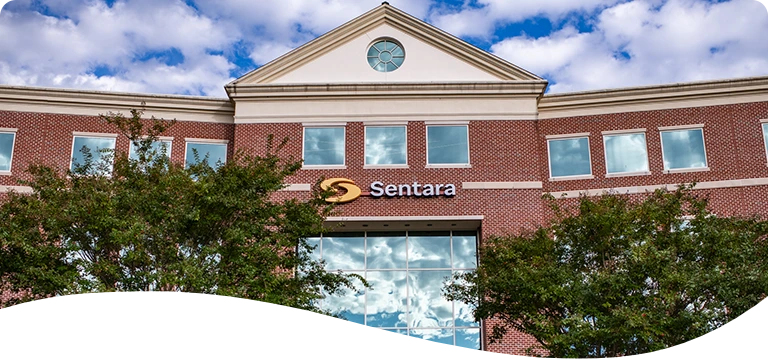 Exterior view of a brick building with large reflective glass windows and the "Sentara" logo displayed above the entrance; colorful balloon decorations flank the doorway, and trees partially frame the scene under a partly cloudy sky.