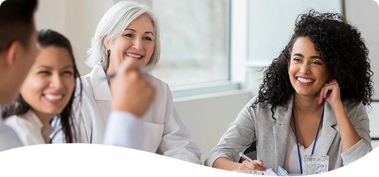 A group of professionals, including women in business attire and medical uniforms, sit around a conference table engaged in a cheerful and collaborative meeting in a bright, modern office setting.