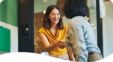 A cheerful woman in a mustard-yellow top smiles while shaking hands with another person in an indoor office setting, suggesting a friendly greeting or successful meeting.