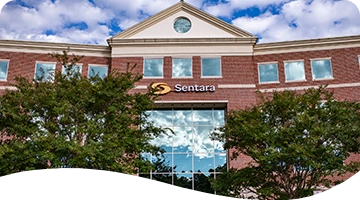 Exterior view of a brick building with large reflective glass windows and the "Sentara" logo displayed above the entrance; colorful balloon decorations flank the doorway, and trees partially frame the scene under a partly cloudy sky.