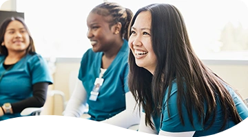 Smiling healthcare workers wearing teal-colored scrubs sit together in a bright room