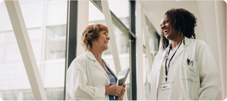 Two female doctors in lab coats smiling and chatting in a hospital hallway.