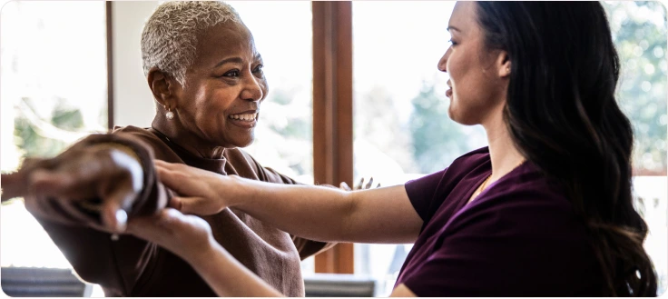 A woman smiling as a healthcare worker helps her with arm exercises in a bright, sunlit room.