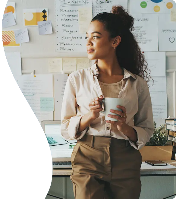 A woman stands in front of a desk holding a coffee mug, looking thoughtfully to the side; she is surrounded by notes, charts, and planning materials pinned to the wall behind her, suggesting a creative or project-focused workspace.