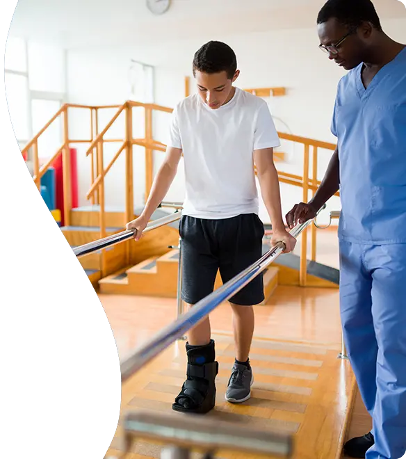 A young man wearing a medical walking boot practices walking with the support of parallel bars while a healthcare professional in scrubs closely assists him in a physical therapy setting.