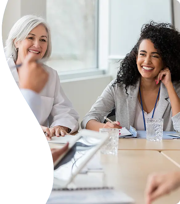 A group of professionals, including women in business attire and medical uniforms, sit around a conference table engaged in a cheerful and collaborative meeting in a bright, modern office setting.