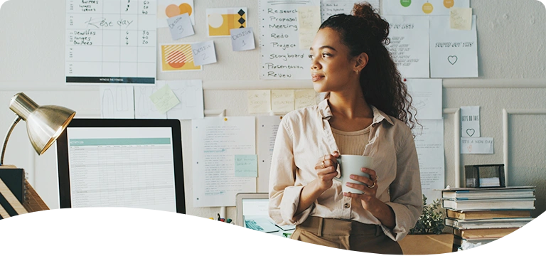 A woman stands in front of a desk holding a coffee mug, looking thoughtfully to the side; she is surrounded by notes, charts, and planning materials pinned to the wall behind her, suggesting a creative or project-focused workspace.