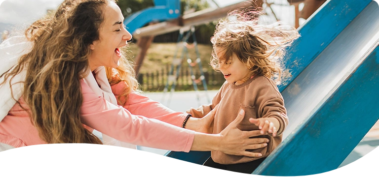A joyful woman greets a laughing child at the bottom of a playground slide on a sunny day, both sharing a moment of happiness and connection.