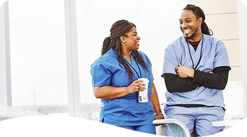 Two healthcare professionals in blue scrubs share a lighthearted conversation and smiles during a break, with one holding a coffee cup in a casual, relaxed setting.