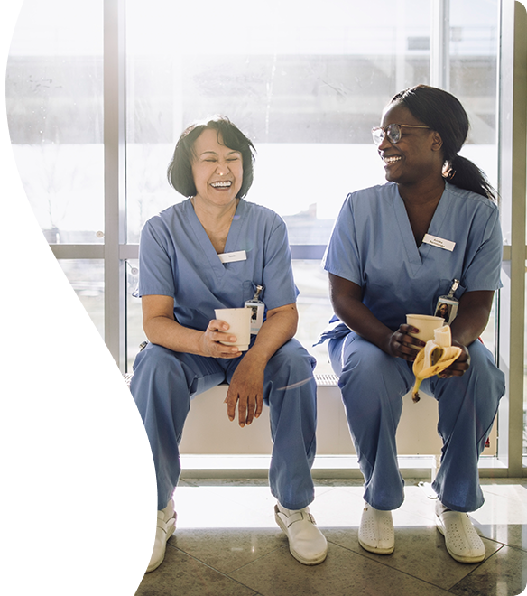 Two female nurses in scrubs laughing and enjoying a break with coffee and a banana in a sunshine filled office with glass wall behind them.