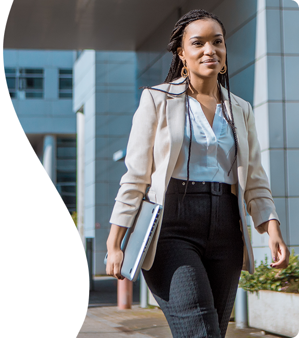 A woman dressed in business attire walking outside an office building with a laptop in hand.