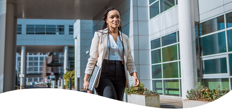 A woman dressed in business attire walking outside an office building with a laptop in hand.