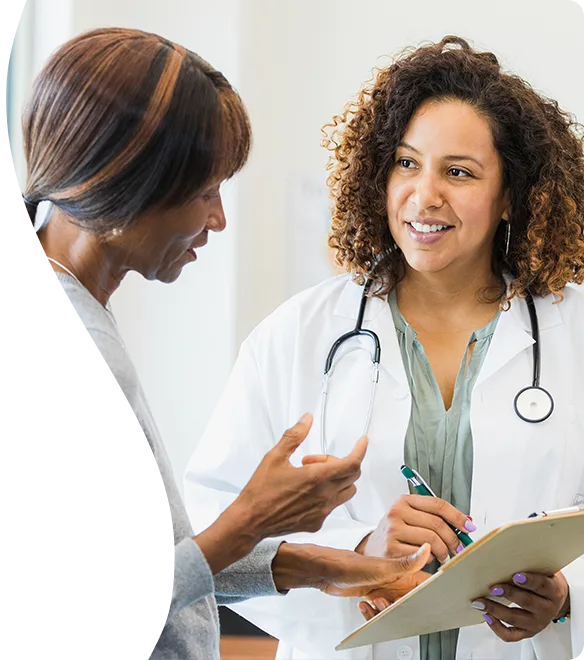 A smiling healthcare worker with a stethoscope around her neck taking notes on a clipboard while talking to another person.