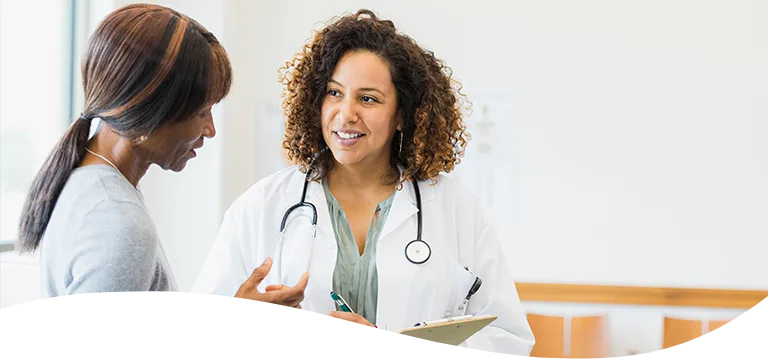 A smiling healthcare worker with a stethoscope around her neck taking notes on a clipboard while talking to another person.