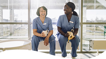 Two female nurses in scrubs laughing and enjoying a break with coffee and a banana in a sunshine filled office with glass wall behind them.