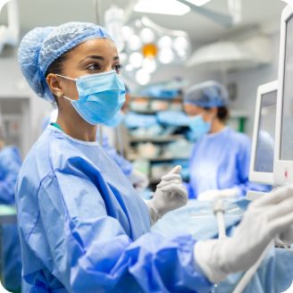 Surgical assistant in scrubs and mask operates medical monitor in an operating room with other staff in the background.