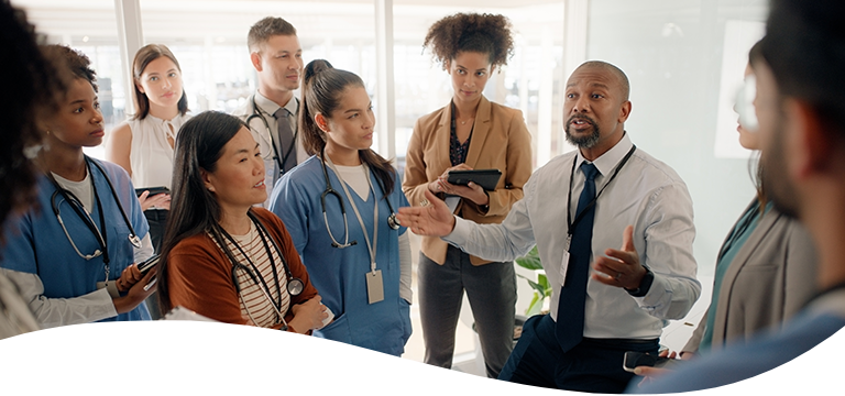 A man in business attire speaks to a group of healthcare professionals, some in scrubs and others in business clothing, gathered closely in a bright office setting.